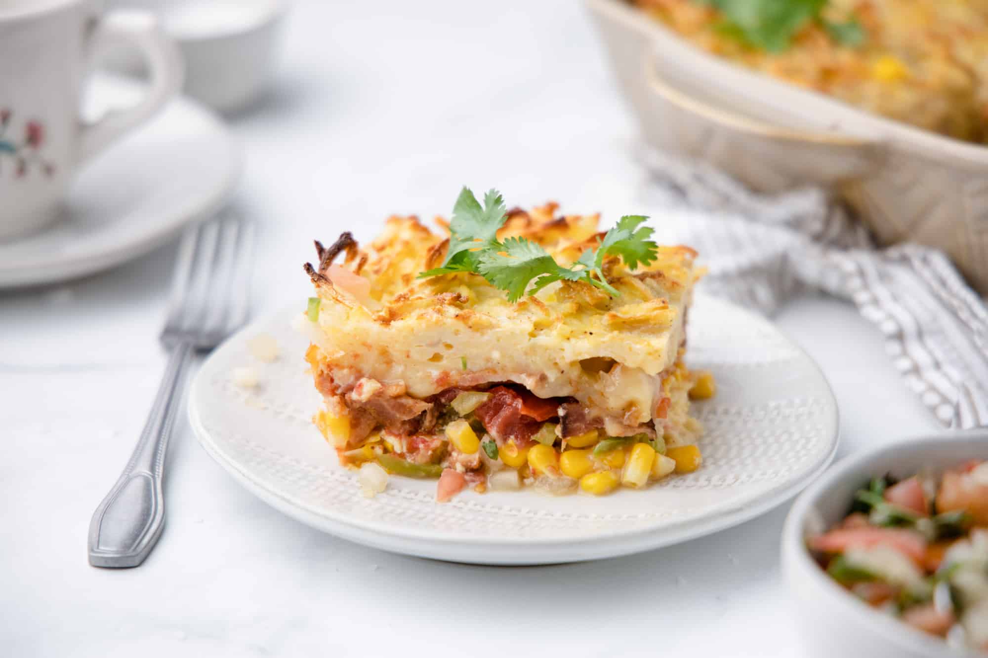 A piece of casserole with corn, bacon and shredded potatoes on a white plate with a fork and salsa.