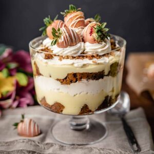 A trifle dish filled with homemade carrot cake, cream cheese whip and vanilla pudding topped with more whipped cream and dipped strawberries