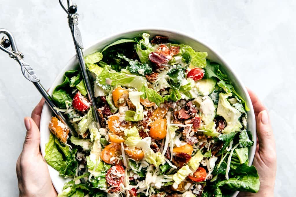 A large green salad with tomatoes, avocado, cheese and bacon in a large bowl placed on a marble countertop, tossed tossed with the tongs on the left