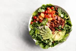 A large green salad with tomatoes, avocado, cheese and bacon in a large bowl placed on a marble countertop