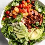 A large green salad with tomatoes, avocado, cheese and bacon in a large bowl