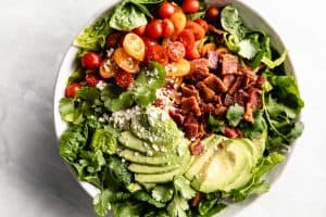 A large green salad with tomatoes, avocado, cheese and bacon in a large bowl