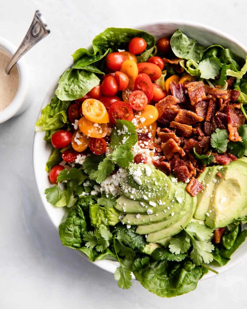 A large green salad with tomatoes, avocado, cheese and bacon in a large bowl placed on a marble countertop, dressing on the side