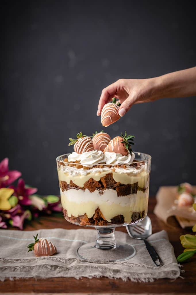 A trifle dish filled with homemade carrot cake, cream cheese whip and vanilla pudding topped with more whipped cream and dipped strawberries