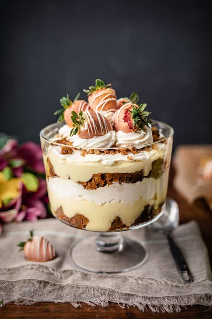 A trifle dish filled with homemade carrot cake, cream cheese whip and vanilla pudding topped with more whipped cream and dipped strawberries