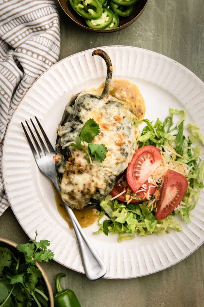 A white plate with a stuffed poblano pepper topped with melted cheese with a side salad of lettuce and tomato