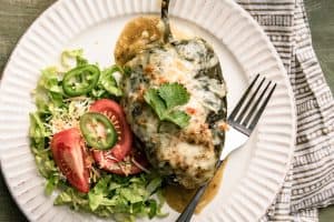 A white plate with a stuffed poblano pepper topped with melted cheese with a side salad of lettuce and tomato