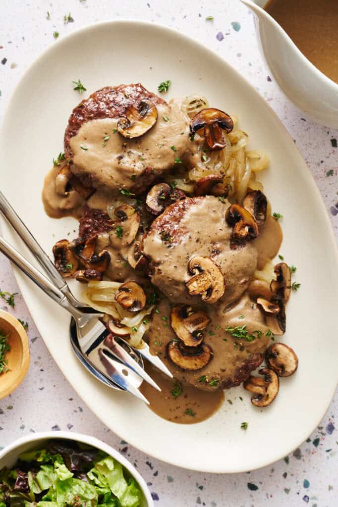 Overhead view of a platter with Oven Hamburger Steaks smothered in mushroom gravy with onions and mushrooms scattered around.