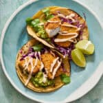 fish tacos on a plate with corn tortillas, limes and guacamole