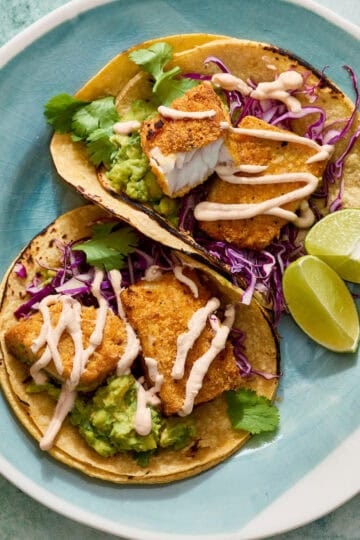fish tacos on a plate with corn tortillas, limes and guacamole