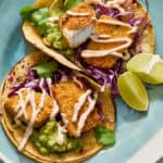 fish tacos on a plate with corn tortillas, limes and guacamole