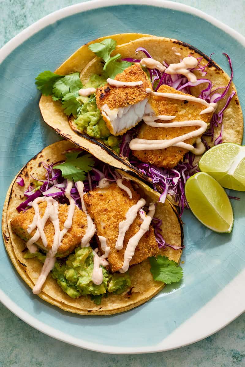 fish tacos on a plate with corn tortillas, limes and guacamole